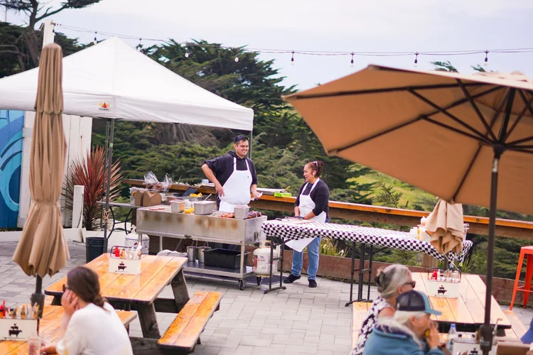 People at an outdoor food stand with grills and tables, surrounded by trees and umbrellas.
