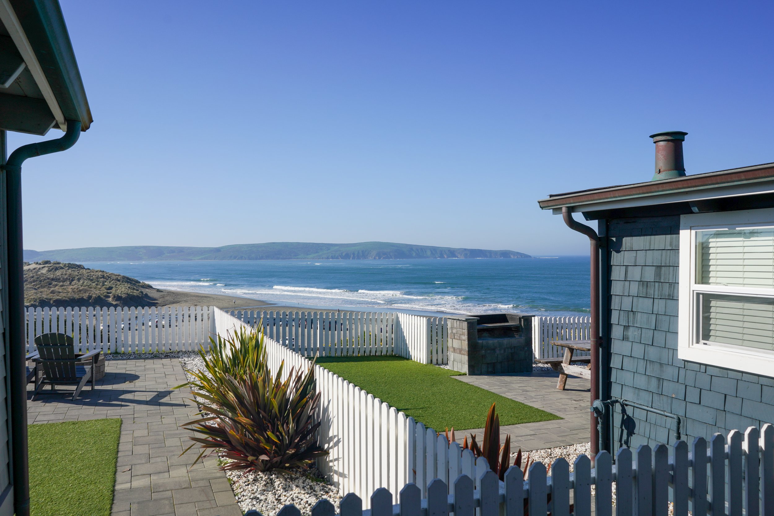 Looking between two cedar-shingle beach houses toward a white picket fence and a panoramic view of the 海岸線 and ocean waves.
