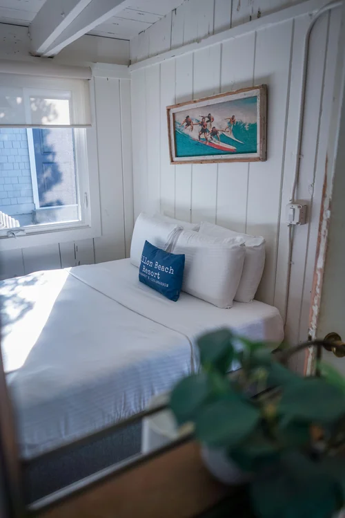 A cozy bedroom with white vertical shiplap walls, a bed with crisp white linens, a navy blue decorative pillow, and vintage-style surf art hanging above the headboard.