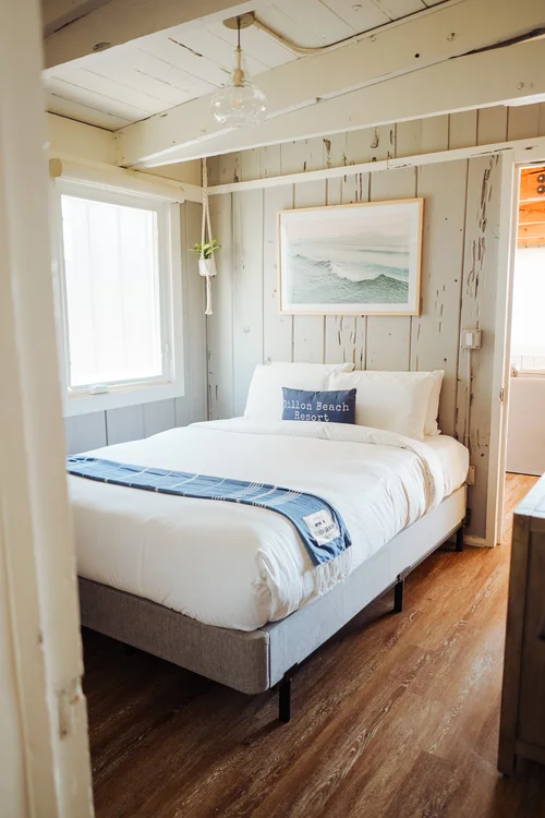 A bright and airy coastal-style bedroom with distressed grey shiplap paneling on the wall, exposed white painted ceiling beams, a clear glass pendant light, and a Queen bed with a navy blue and white beach-themed pillow and ocean wave art above it.