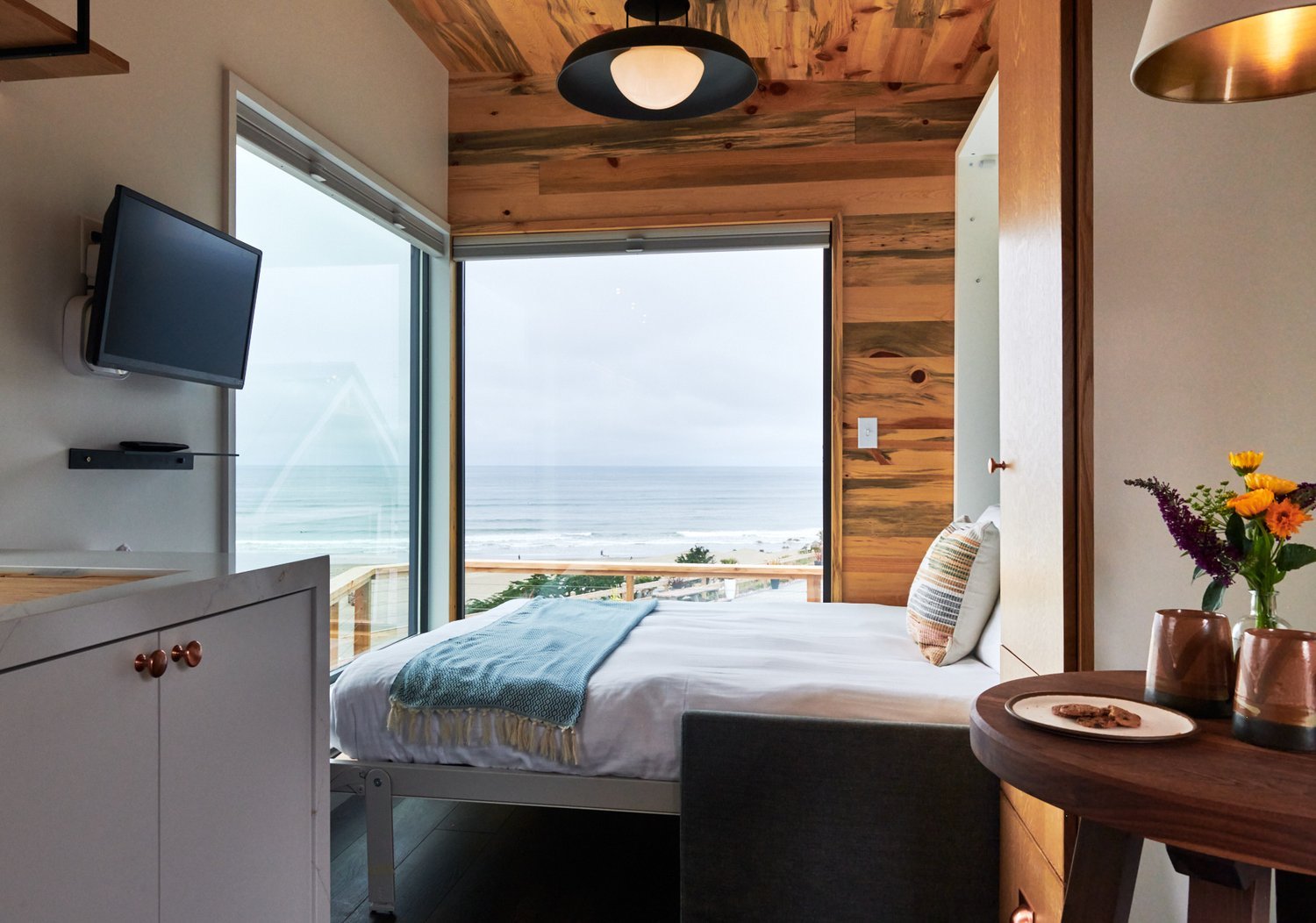 Master bedroom in an Osprey cabin with white shiplap walls, a bed with a blue striped throw, and a window looking out toward the Pacific Ocean.