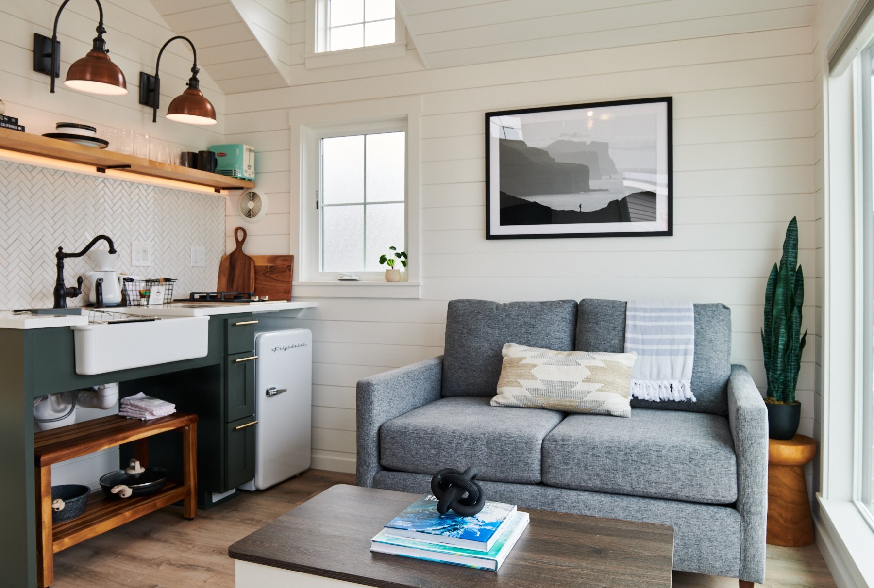 Bright living area with a grey sofa and a large floor-to-ceiling window showcasing a coastal landscape and outdoor patio.