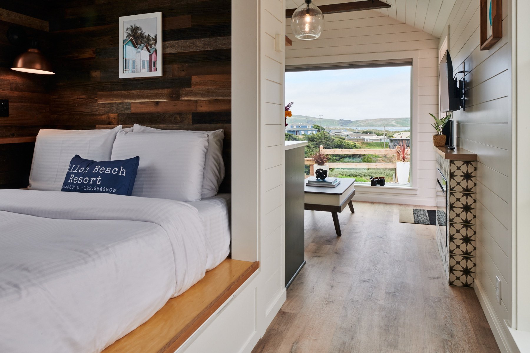 Open-concept tiny house interior showing the relationship between the kitchen, grey sofa, and large viewing window.
