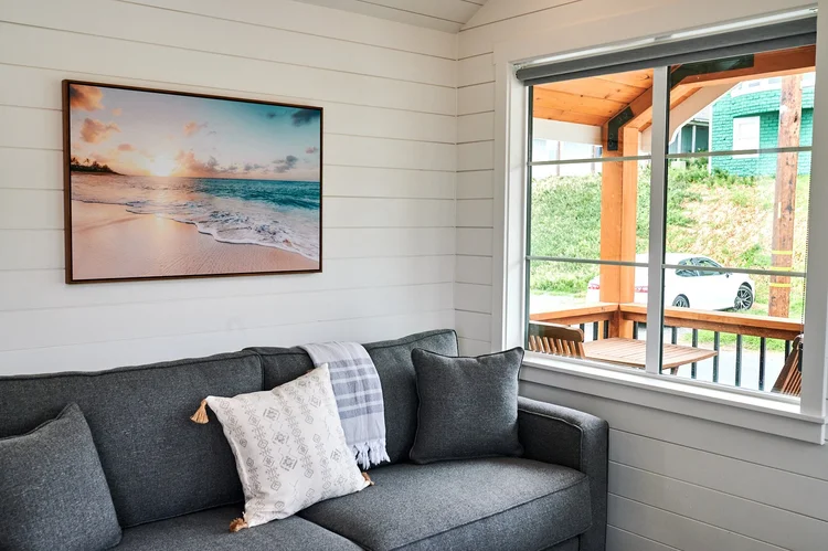 A bright and modern living room corner in a Sandpiper cabin, featuring a grey sofa against a shiplap wall, a large framed canvas of a tropical beach, and a window looking out onto a wooden deck and coastal vegetation.