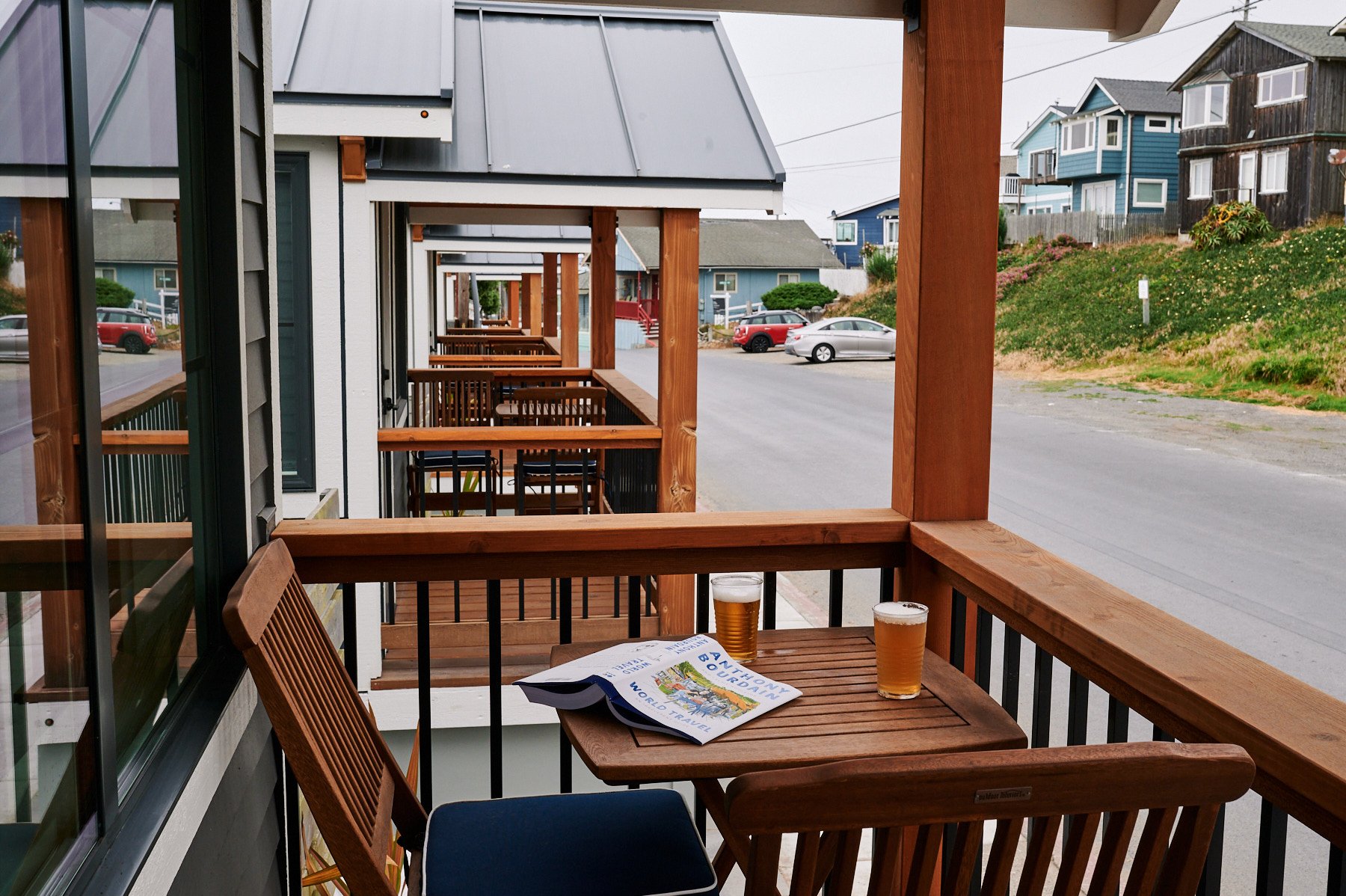 Private wooden deck of a Sandpiper cabin featuring a bistro table with two beers and an Anthony Bourdain book, overlooking a row of similar modern cottages