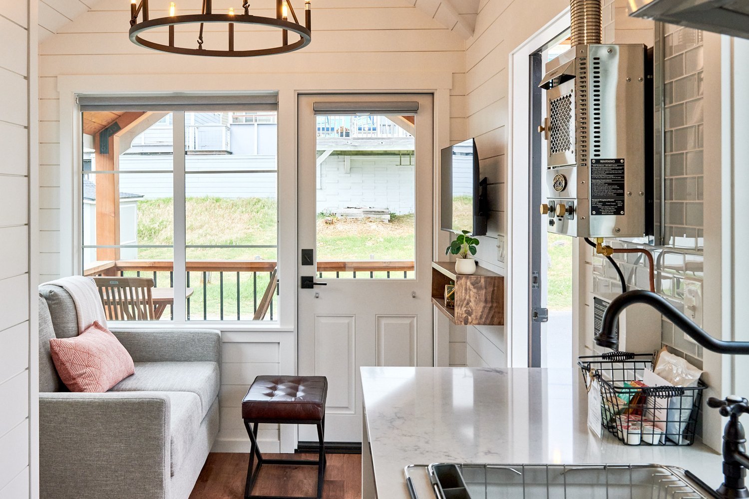 Cozy tiny house interior showing a grey sofa, a leather ottoman, and a large window overlooking the porch.  