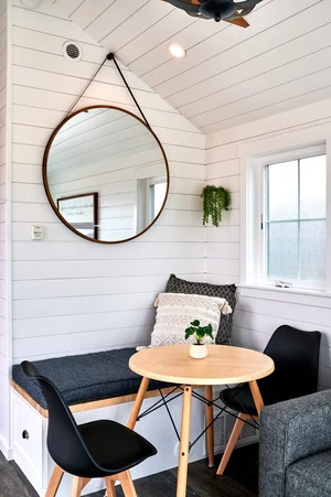 Modern interior of a Sandpiper cabin nook featuring a built-in white shiplap banquette with a blue cushion, an adjustable wooden round table, two black mid-century modern chairs, and a large round mirror hanging above.
