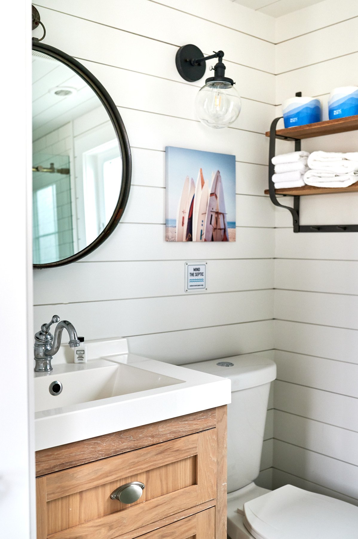 A view into the Sandpiper cabin bathroom showing a light wood vanity with a large white ceramic sink, a chrome faucet, a large round mirror, and a wall art piece featuring a row of surfboards.