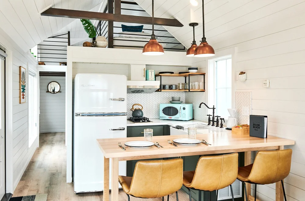 Bright modern interior of a ブライトリビエラ COHO cabin featuring a white retro refrigerator, mint green microwave, and a wooden dining island with three leather stools. A sleeping loft is visible above the kitchen area under a vaulted ceiling wit