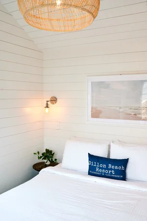 A close-up of the Sandpiper cabin bedroom showing a queen bed with crisp white linens, a framed photograph of a beach sunset, a wicker pendant light, and a custom blue throw pillow embroidered with 'ブライトリビエラ' and coordinates.