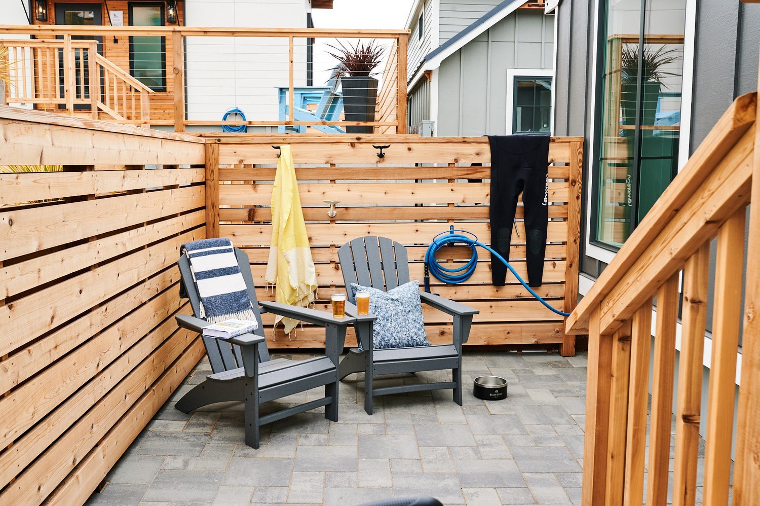 A private, fully-enclosed stone paver patio on the lower level of the Sandpiper cabin, featuring two dark grey Adirondack chairs with patterned pillows and a small table between them, enclosed by a modern horizontal slatted wood fence.