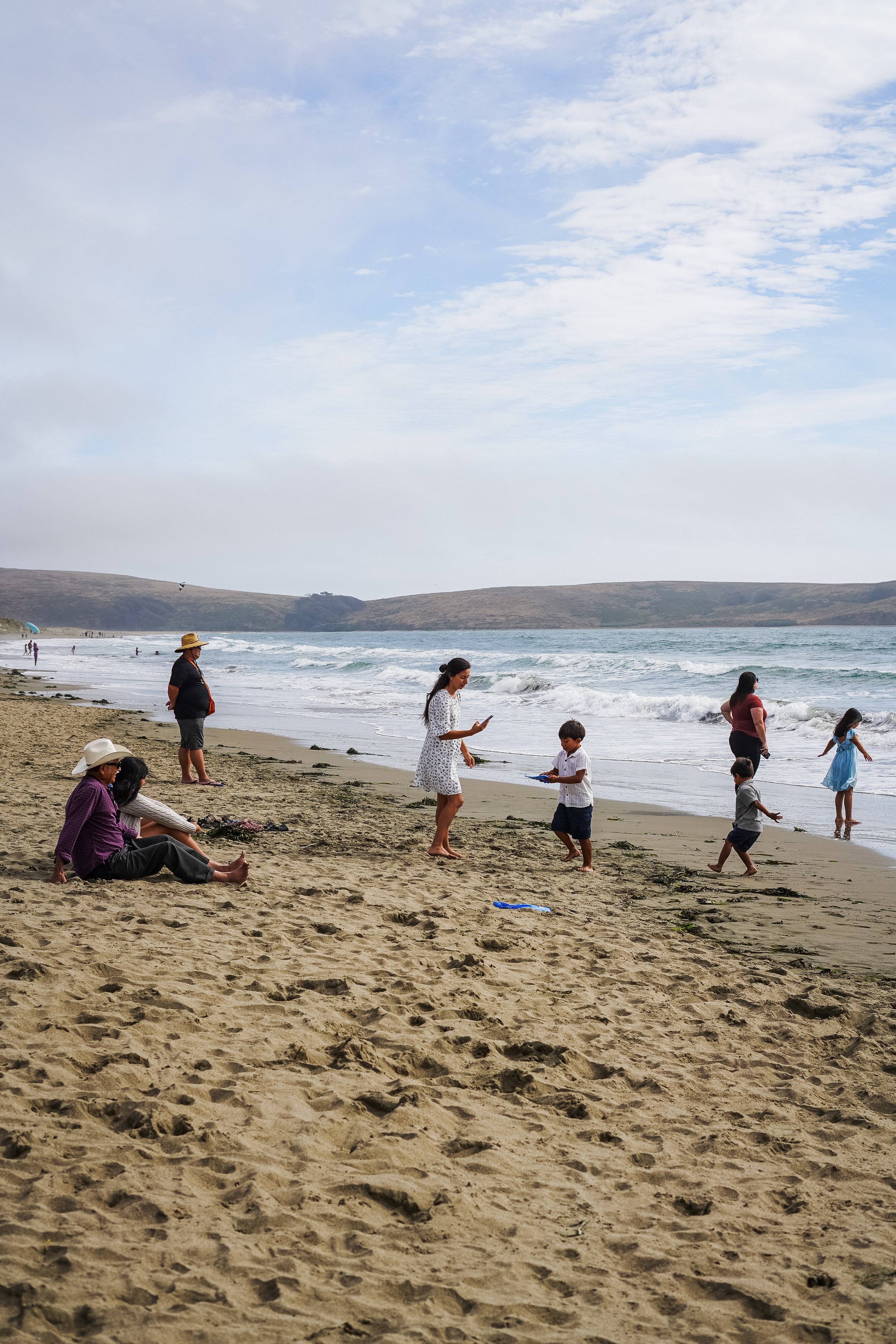 People enjoying a day at the beach; some are sitting on the sand, others are standing or walking near the shoreline with small waves. The sky is partly cloudy with a distant hilly landscape in the background.