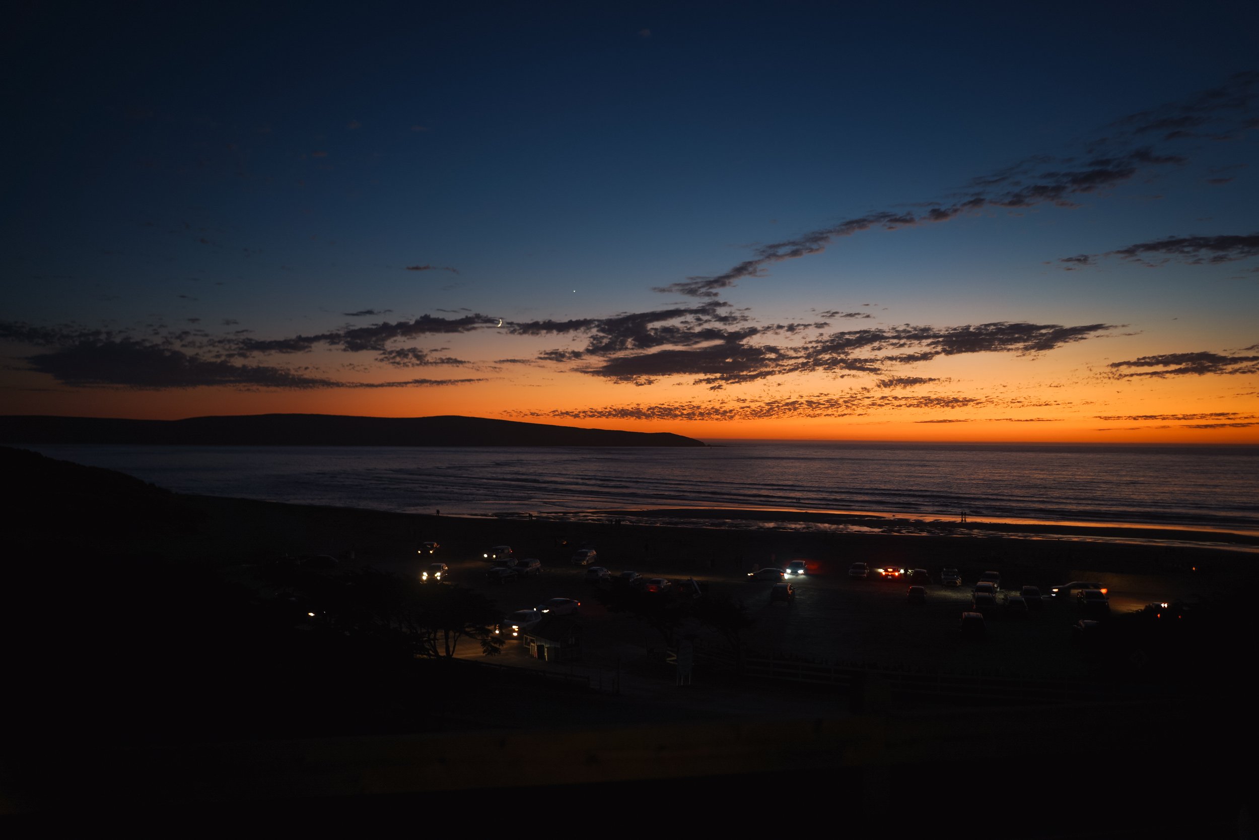 Sunset over the ocean with a dark shoreline, scattered clouds in the sky, and cars parked along a road in the foreground.