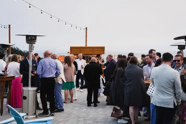 An outdoor gathering with people mingling near a bar and patio heaters, under string lights with a cloudy sky in the background.