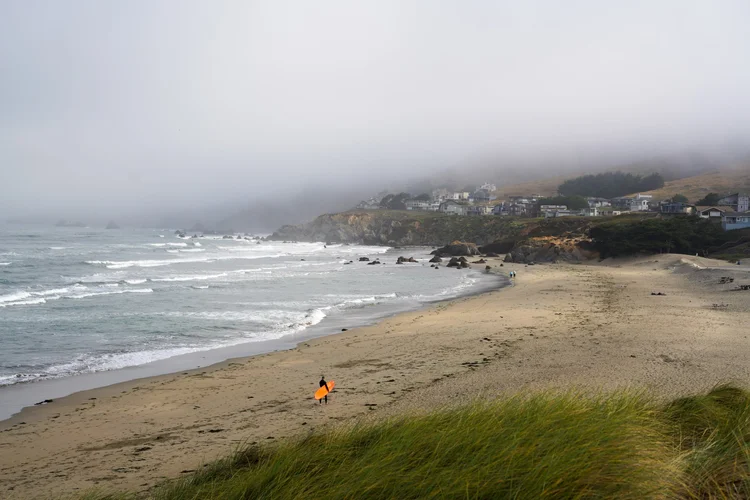A foggy beach scene with houses on a hillside, a person carrying an orange surfboard walking on the sand, and ocean waves crashing against rocks.