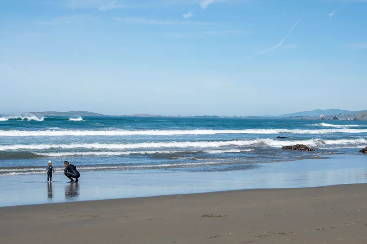A beach with people in wetsuits preparing for surfing, ocean waves, and a distant shoreline with hills under a partly cloudy sky.