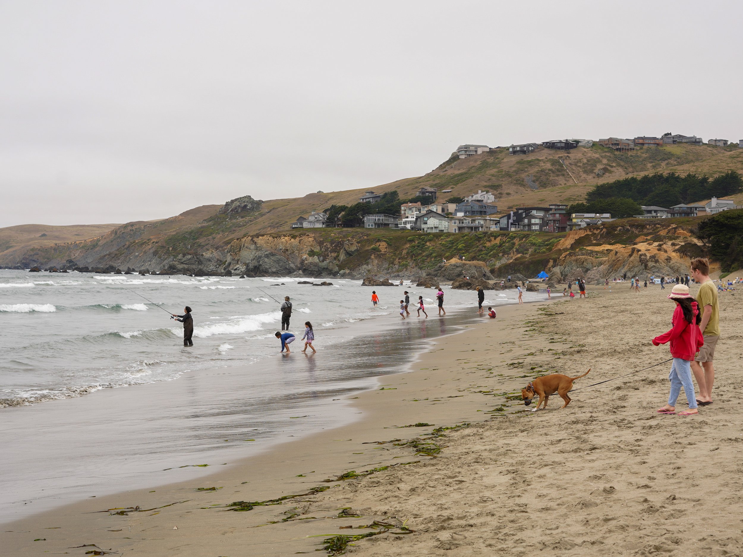 People walking and fishing on a beach with rocky cliffs and houses on a hillside in the background.