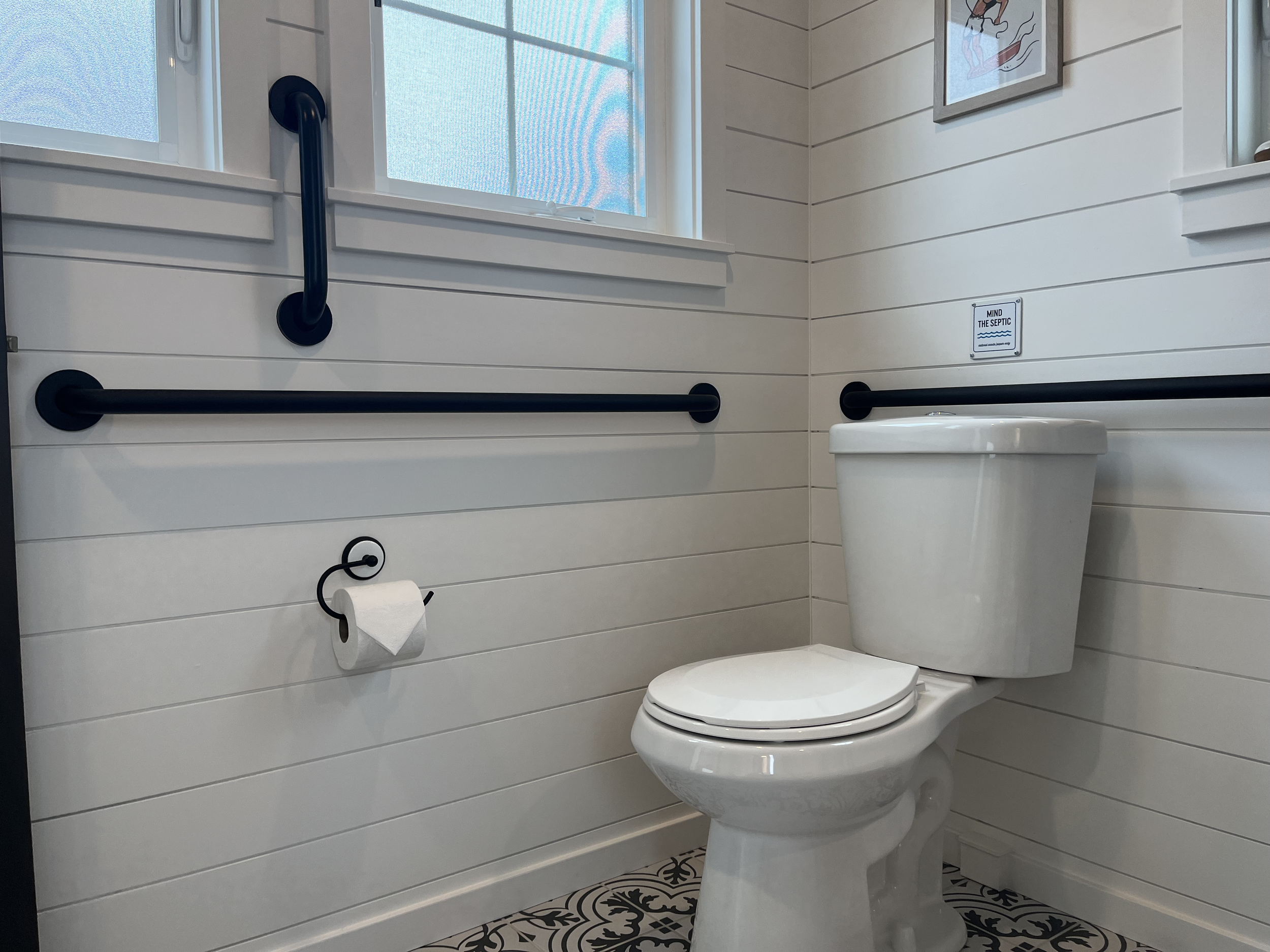 Close-up of an ADA-compliant toilet featuring black matte horizontal and vertical grab bars against white shiplap walls.
