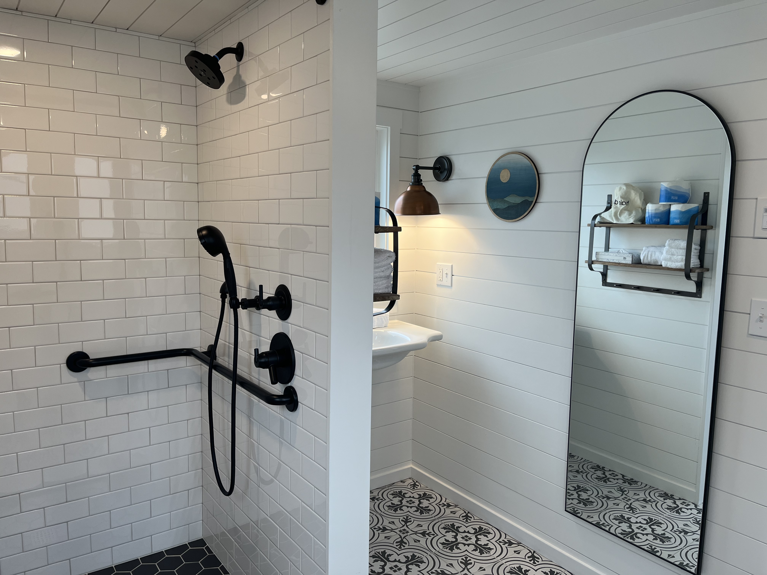 Bathroom interior showing a zero-entry walk-in shower with white subway tile, black fixtures, and a large arched floor mirror.