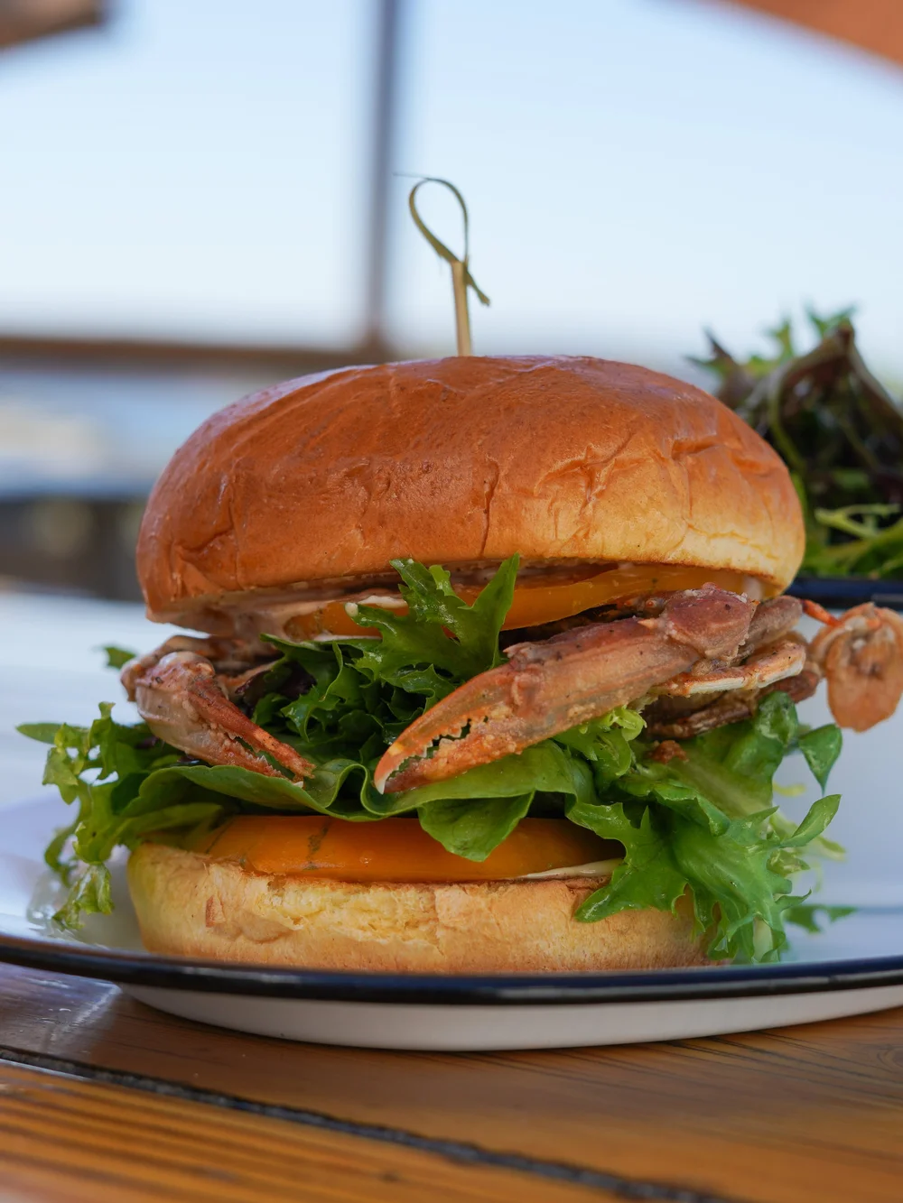A gourmet sandwich on a plate with lettuce, tomato, and bacon, topped with a golden-brown bun held together with a toothpick, placed on a wooden surface.