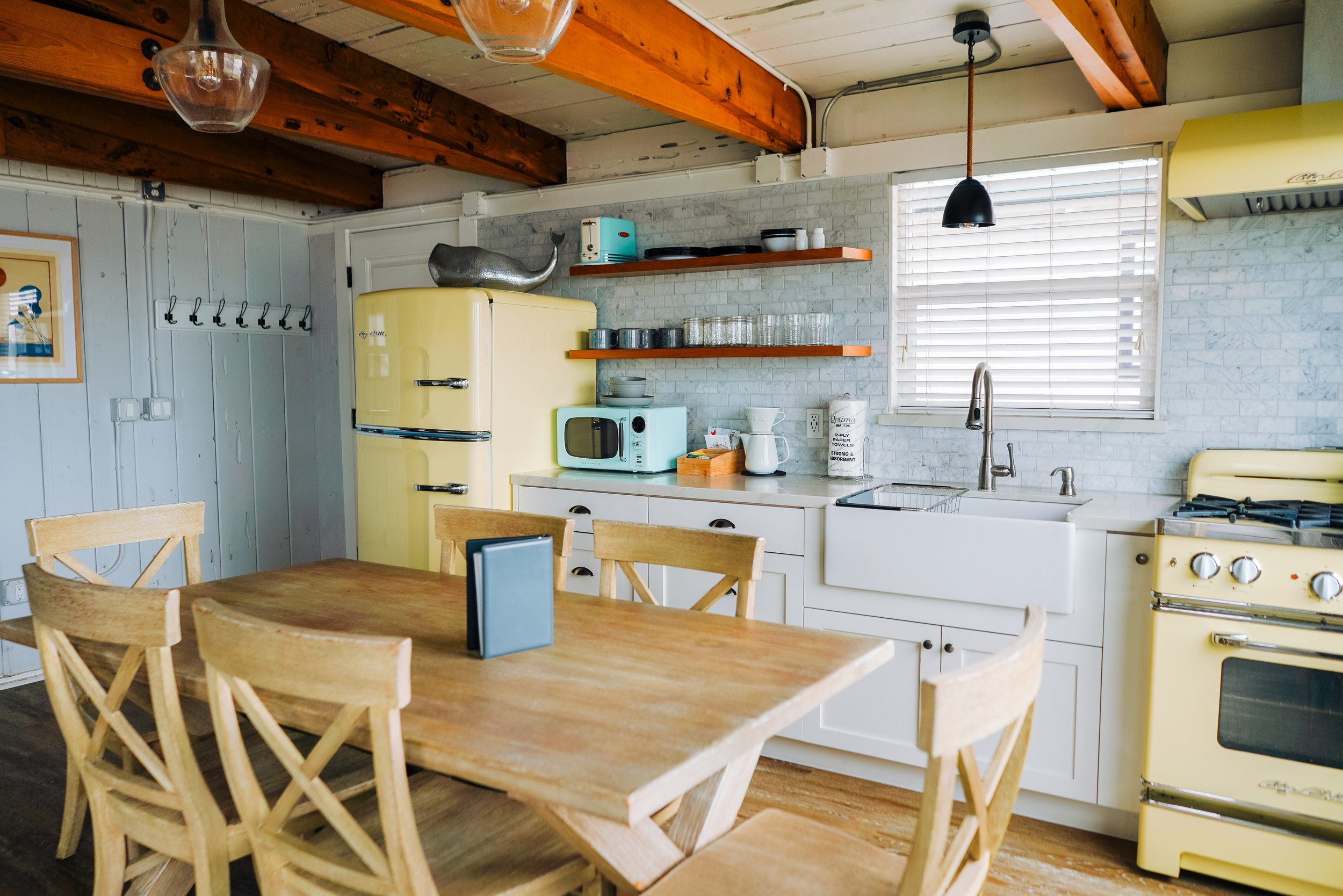 Rustic dining area and kitchen featuring a light wood table, exposed timber ceiling beams, a vintage yellow refrigerator, and mint green appliances against a marble tile backsplash.