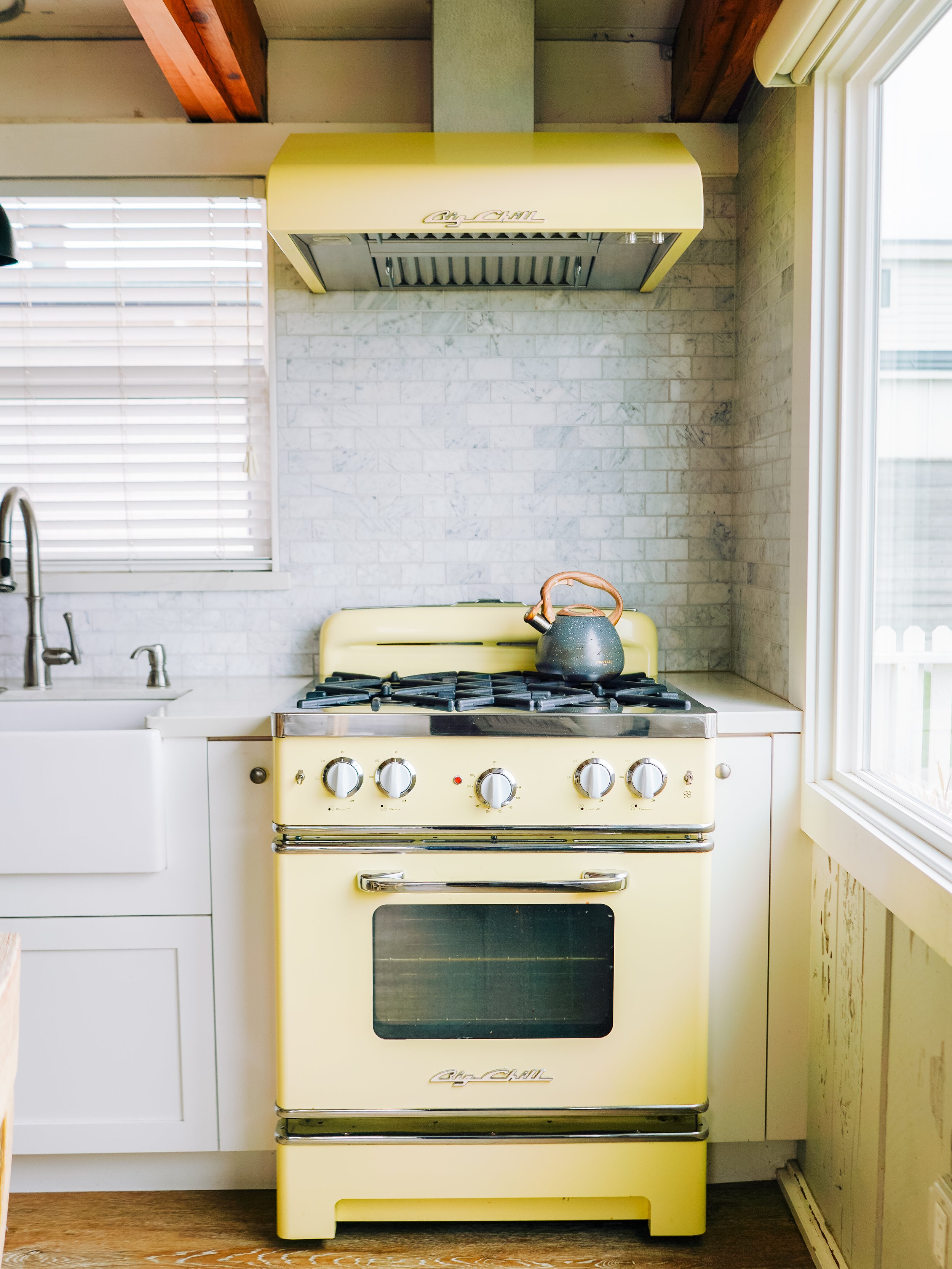 Detail of a pale yellow retro-style Big Chill gas range and matching vent hood against a white marble tile backsplash in a coastal kitchen.