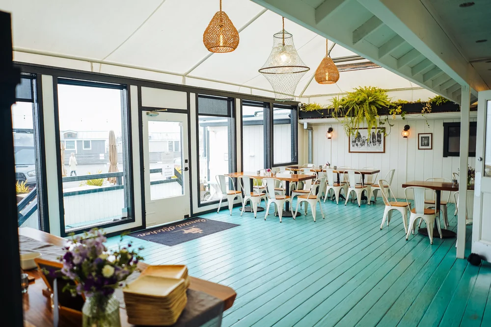 Interior view of a bright sunroom or patio area with large windows, hanging woven light fixtures, green-painted wooden floor, white chairs, framed artwork on the wall, plants, and a table with a vase of flowers.