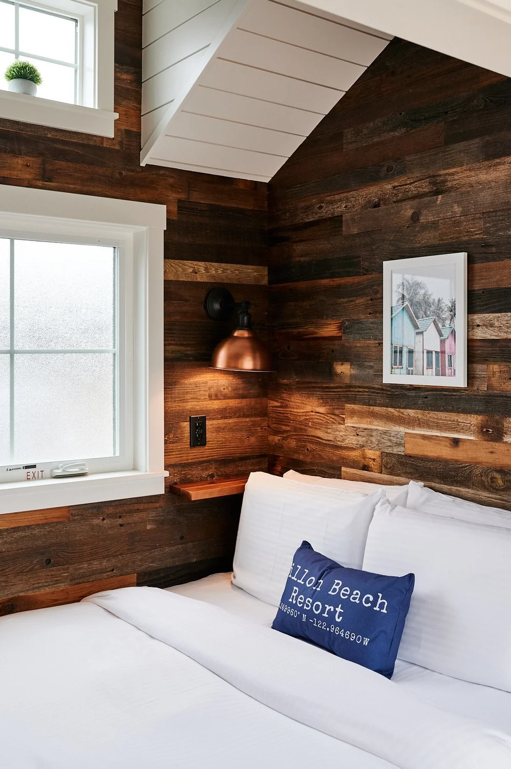 Accessible sleeping nook featuring reclaimed dark wood plank walls, a copper bedside sconce, and white linens with a ブライトリビエラ accent pillow.